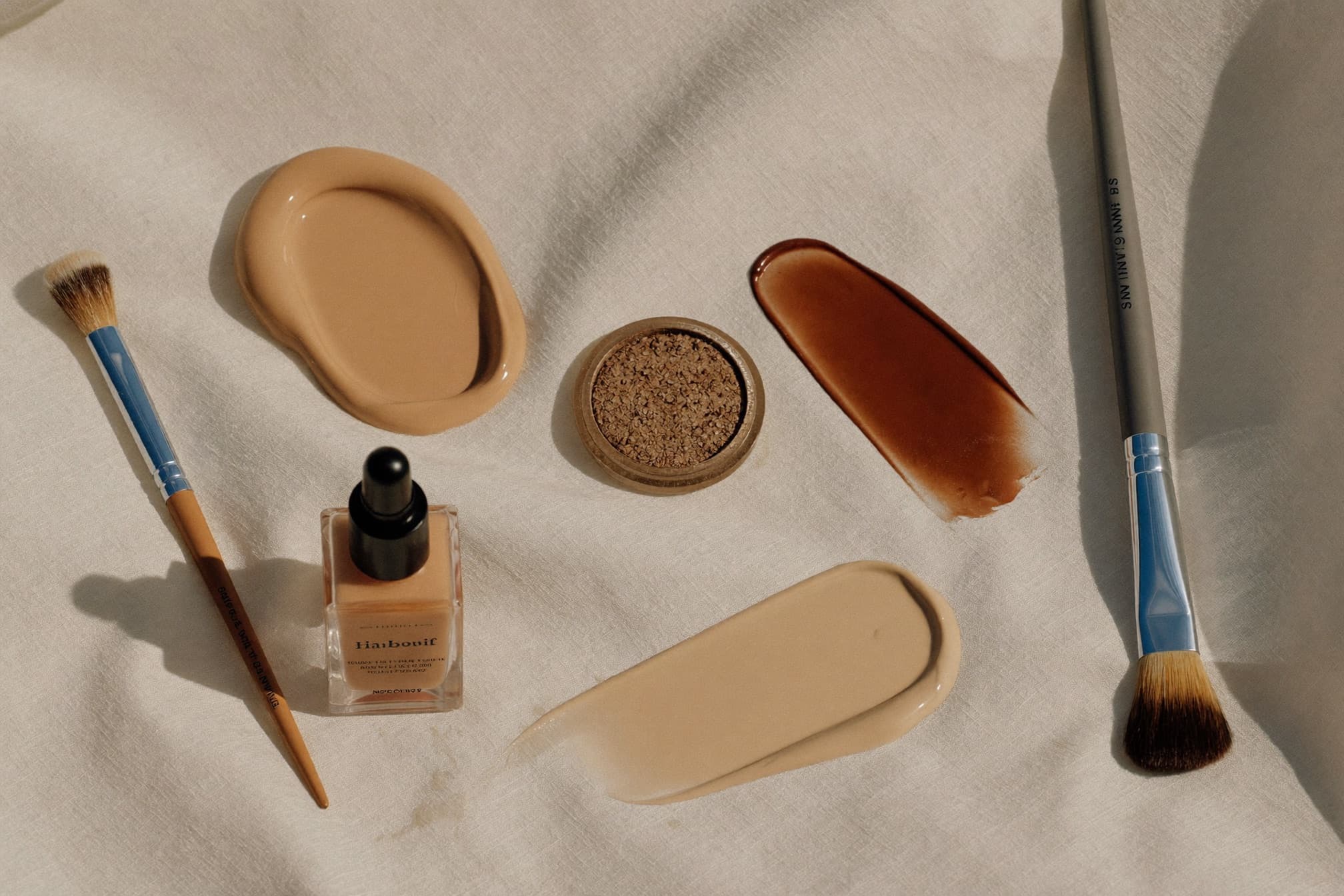 Still life of neutral makeup textures and brushes on ivory linen
